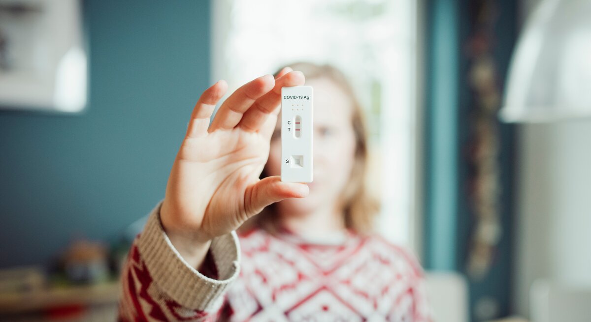 Eine Frau mittleren Alters, die einen rot-weiß-gemusterten Pullover trägt, hält einen positiven Corona-Test in die Kamera, der anzeigt, dass sie die Corona-Mutation Priola hat.