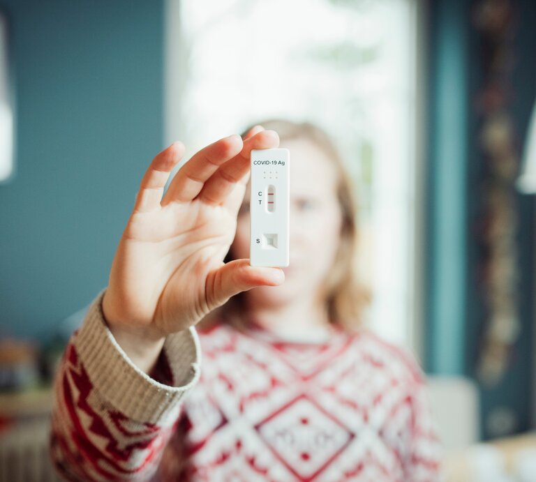 Eine Frau mittleren Alters, die einen rot-weiß-gemusterten Pullover trägt, hält einen positiven Corona-Test in die Kamera, der anzeigt, dass sie die Corona-Mutation Priola hat. | © Adobe Stock/ pascalskwara