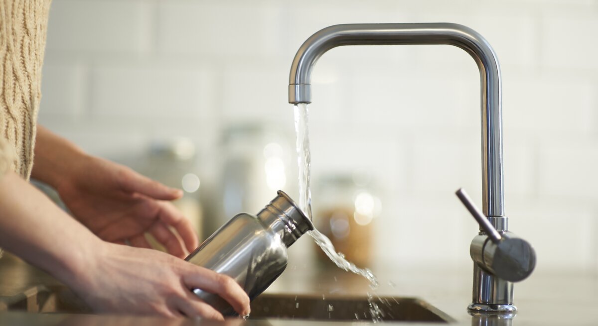 Person füllt eine Wasserflasche aus Edelstahl an einem Wasserhahn auf. Die Szene spielt sich in einer modernen Küche ab, die eine saubere, helle und minimalistische Atmosphäre hat.