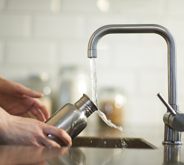 Person füllt eine Wasserflasche aus Edelstahl an einem Wasserhahn auf. Die Szene spielt sich in einer modernen Küche ab, die eine saubere, helle und minimalistische Atmosphäre hat. | © Getty Images/Dougal Waters