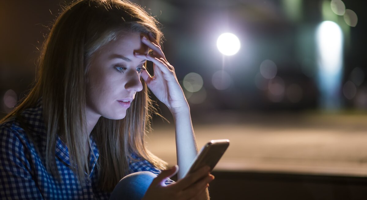 Junge Frau schaut in einem dunklen Zimmer überrascht auf ihr Handy