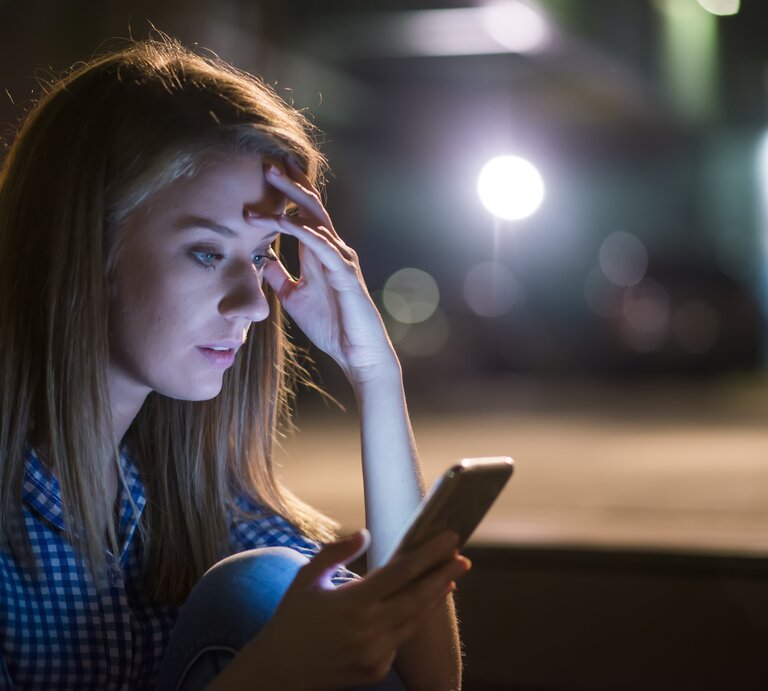 Junge Frau schaut in einem dunklen Zimmer überrascht auf ihr Handy | © iStock.com/dragana991
