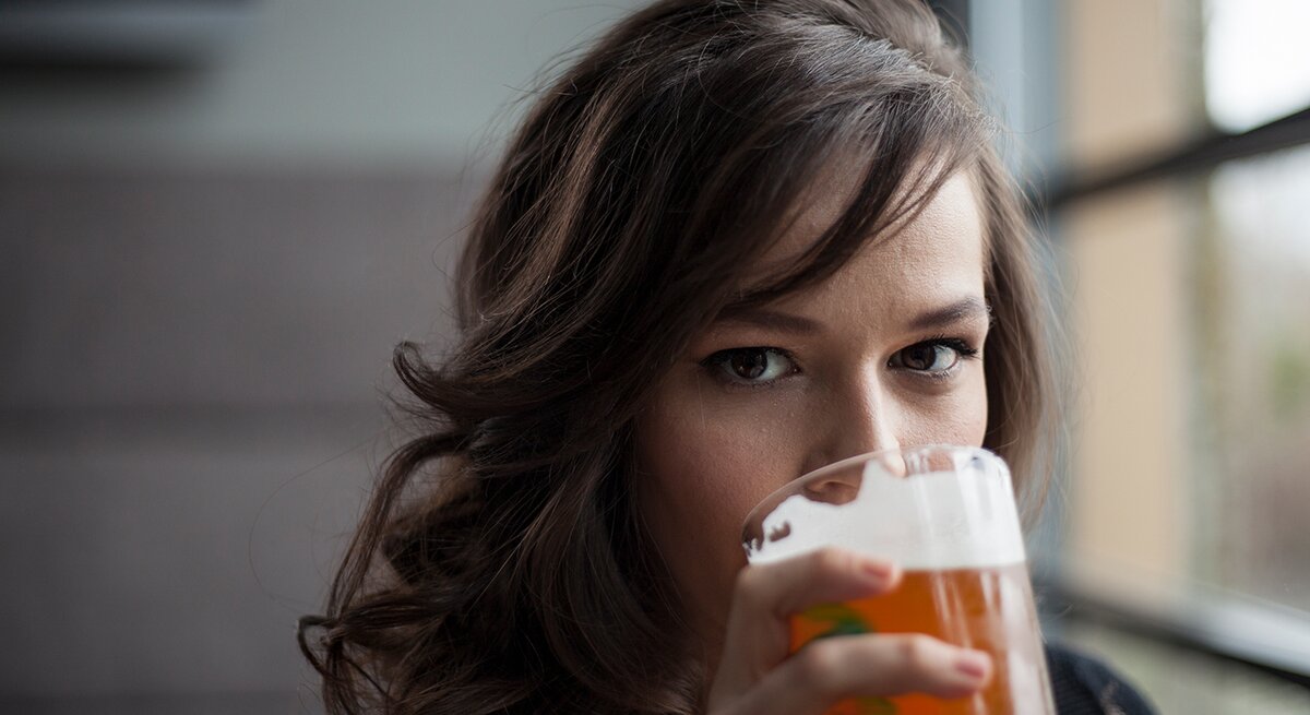 Eine Frau mit lockigem braunem Haar hält ein Glas Bier & trinkt, während sie direkt in die Kamera blickt.