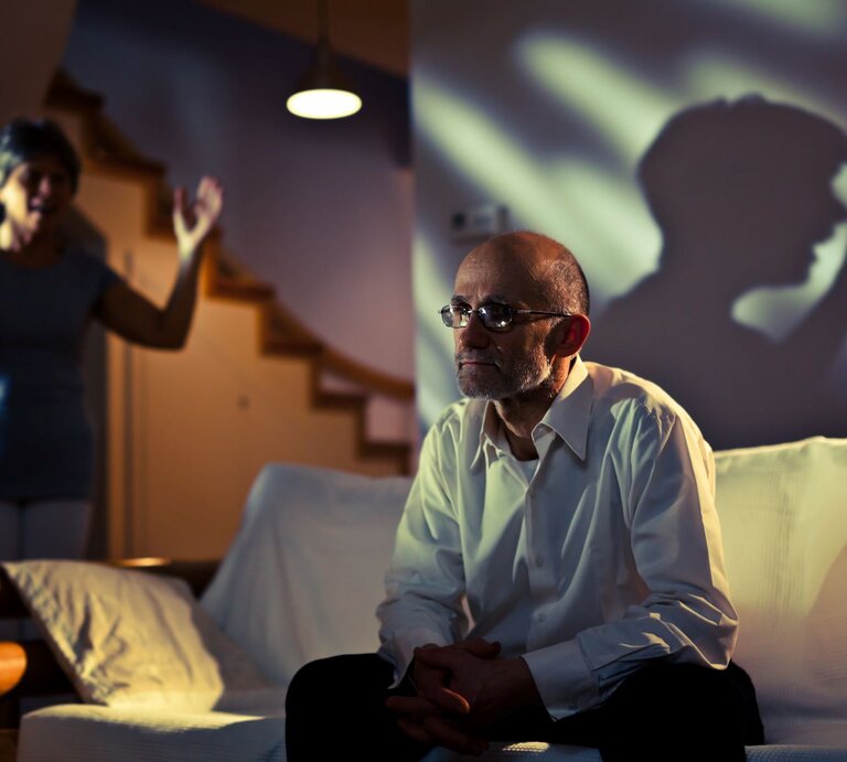 Paar in einem scheinbar intensiven Streit. Der Mann sitzt auf einem Sofa und wirkt nachdenklich und bedrückt, während die Frau im Hintergrund steht und gestikuliert, als ob sie etwas erklärt oder sich beschwert. Die Szene ist dunkel und beleuchtet. | © Getty Images / vm