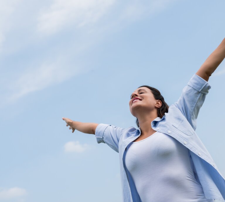  Das Bild zeigt eine Frau, die mit ausgebreiteten Armen nach oben steht und den Himmel anschaut. Sie scheint ein Gefühl von Freiheit oder Erleichterung zu erleben, vielleicht genießt sie einen Moment der Ruhe oder des Triumphes. Die Frau trägt eine blaue Bluse und ein weißes Top, und der Hintergrund besteht aus einem klaren blauen Himmel mit einigen leichten Wolken. Die Pose der Frau und ihr Gesichtsausdruck vermitteln ein starkes Gefühl der Freude und der Offenheit gegenüber der Welt um sie herum. | © iStock/DragonImages