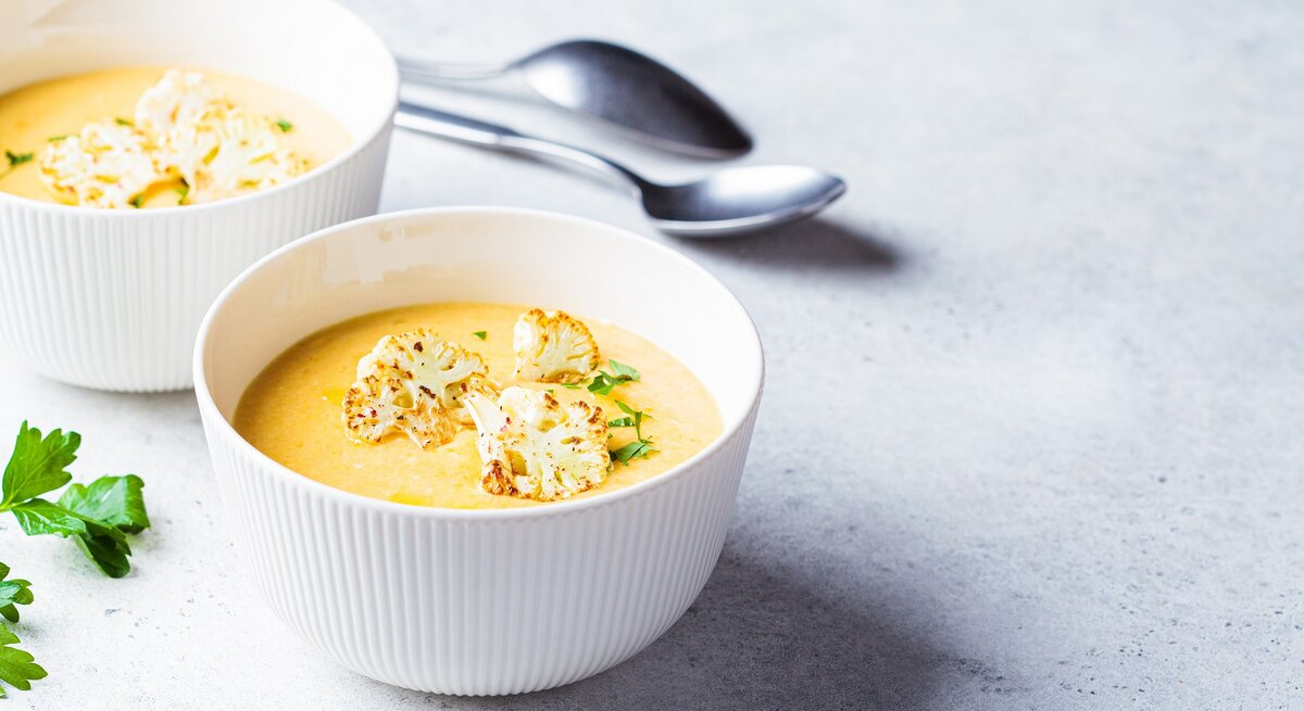 Zwei Schüsseln Blumenkohl-Kartoffel-Suppe mit Kräutern und gerösteten Blumenkohlröschen garniert, dahinter zwei Löffel.