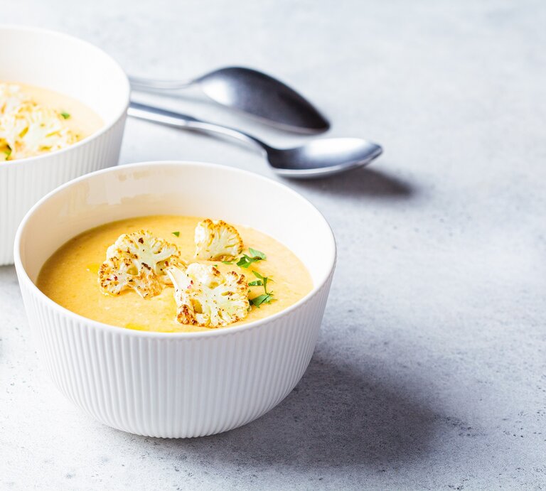 Zwei Schüsseln Blumenkohl-Kartoffel-Suppe mit Kräutern und gerösteten Blumenkohlröschen garniert, dahinter zwei Löffel. | © Adobe Stock/ vaaseenaa