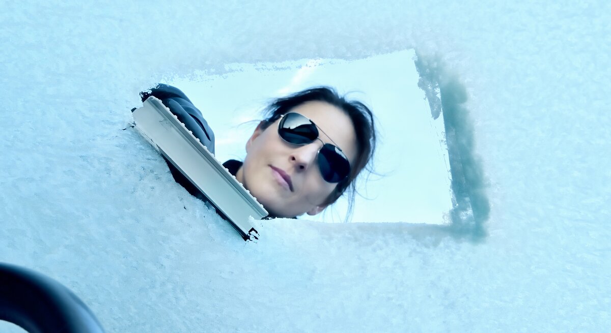 Blick aus dem Inneren durch die Frontscheibe eines Autor, während eine Frau den Schnee entfernt. 