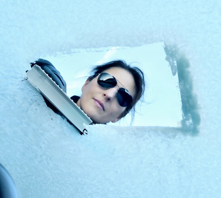 Blick aus dem Inneren durch die Frontscheibe eines Autor, während eine Frau den Schnee entfernt.  | © iStock/trendobjects