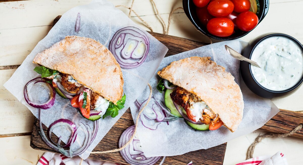 Pita-Gyros mit Beilagen auf Küchenkrepp auf hellem Untergrund. Daneben Schale mit Tzaziki und kleine Tomaten. Draufsicht.