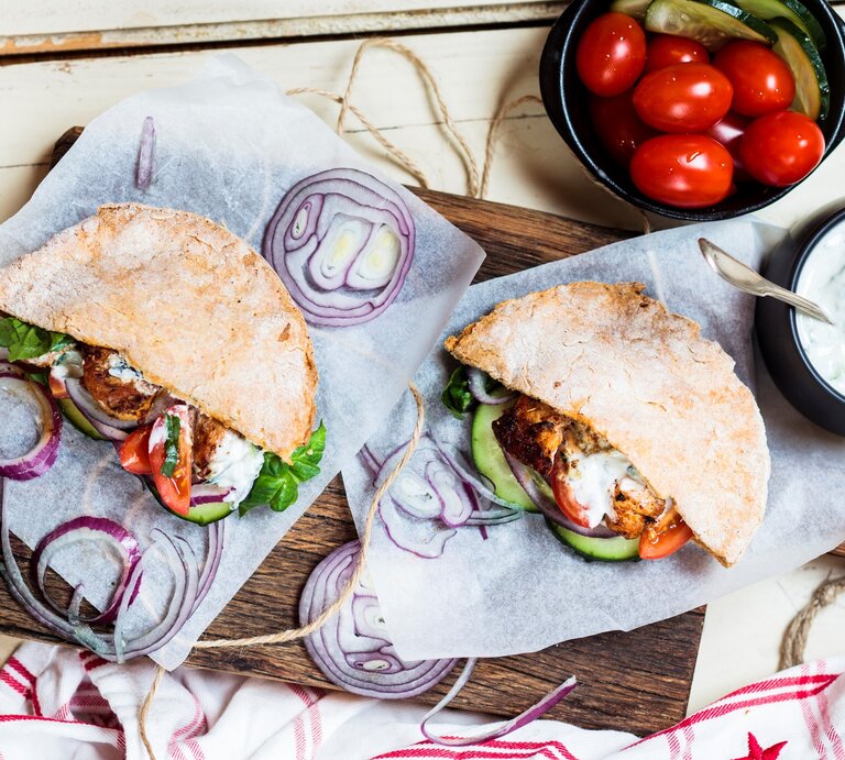 Pita-Gyros mit Beilagen auf Küchenkrepp auf hellem Untergrund. Daneben Schale mit Tzaziki und kleine Tomaten. Draufsicht. | © Getty Images / Westend61