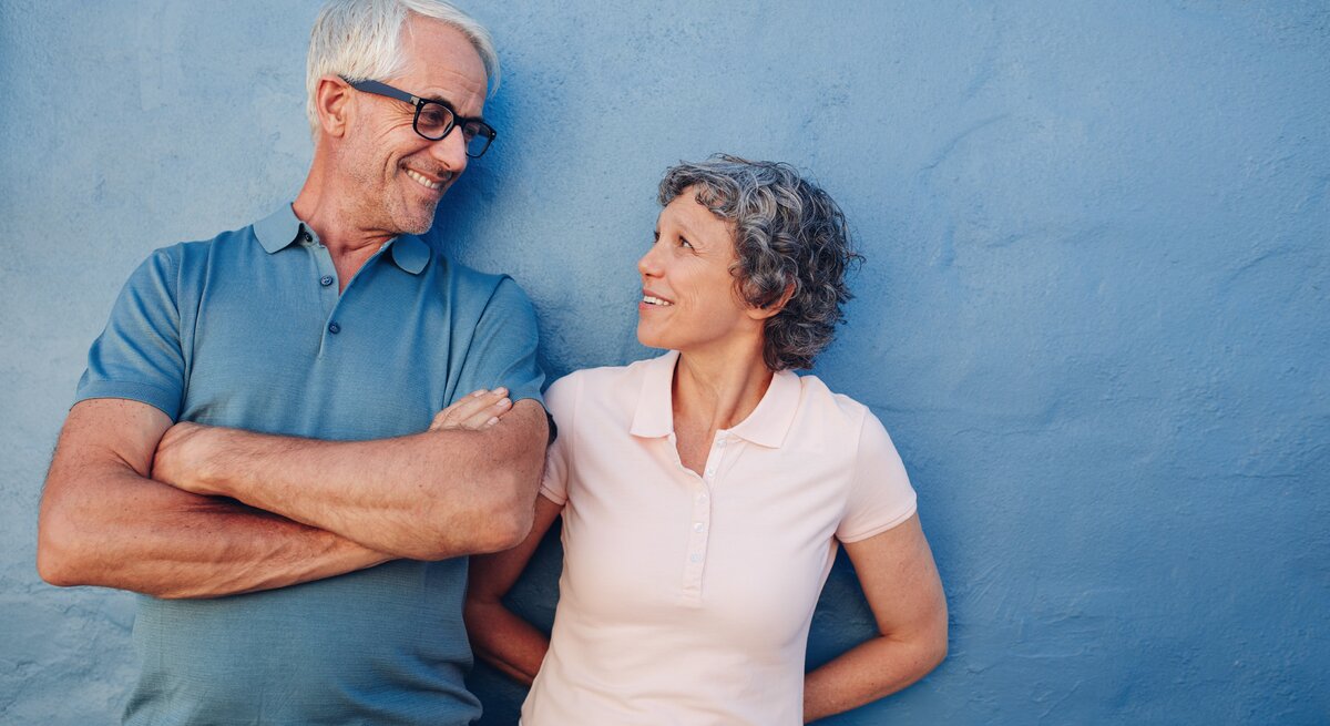 Ein älteres Paar mit grauem Haar steht lachend vor einer blauen Wand, der Mann trägt ein blaues Poloshirt, die Frau ein rosafarbenes.