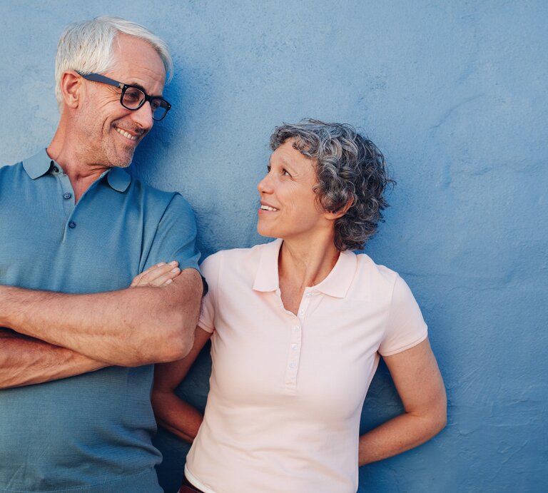 Ein älteres Paar mit grauem Haar steht lachend vor einer blauen Wand, der Mann trägt ein blaues Poloshirt, die Frau ein rosafarbenes. | © iStock.com/jacoblund