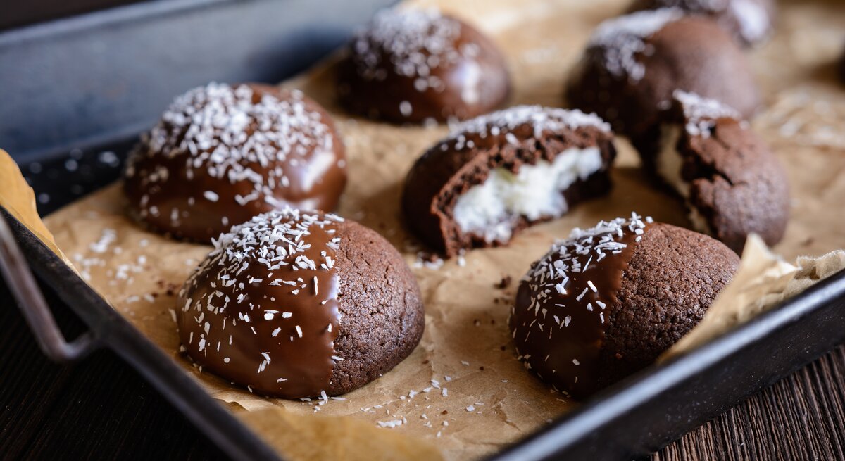Schokocookies mit Kokosfüllung, Schokoglasur und Kokosflocken auf einem Backblech mit Backpapier auf einem dunklen Holztisch. 