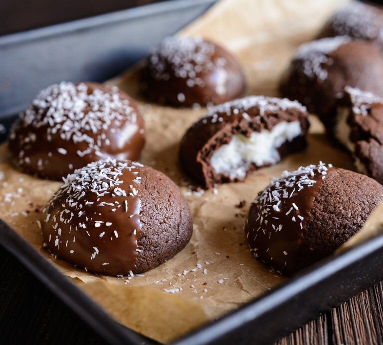 Schokocookies mit Kokosfüllung, Schokoglasur und Kokosflocken auf einem Backblech mit Backpapier auf einem dunklen Holztisch.  | © Getty Images / NoirChocolate