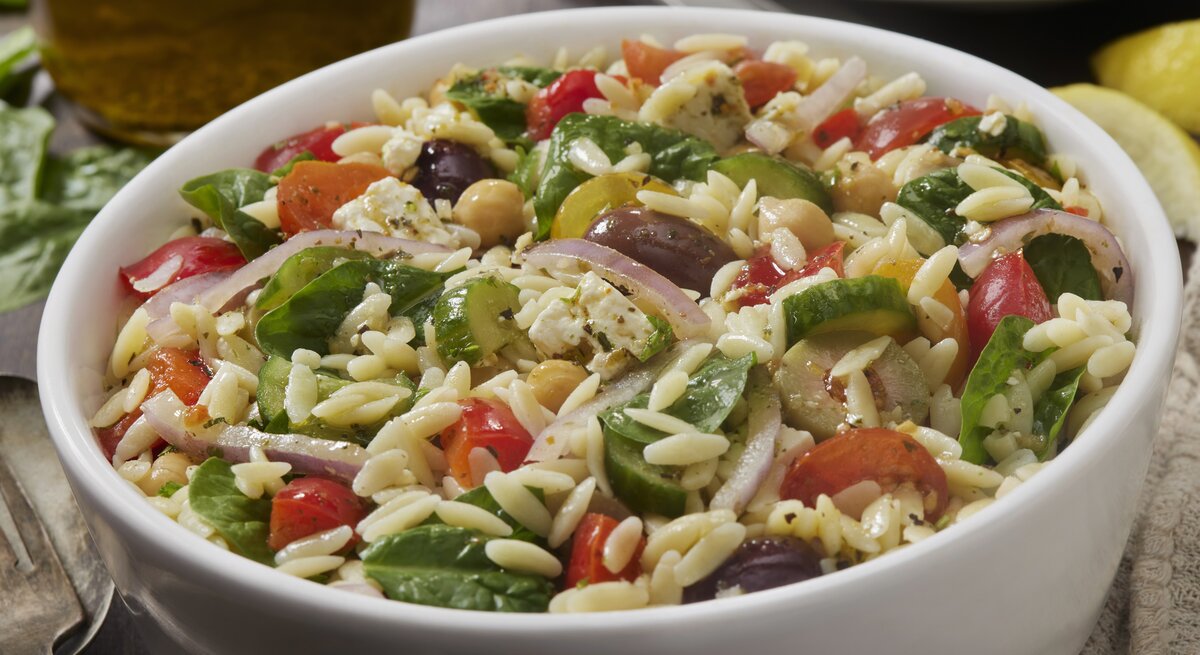 Mediterraner Orzo-Salat mit Spinat, Tomaten und Oliven in weißer Schüssel. 