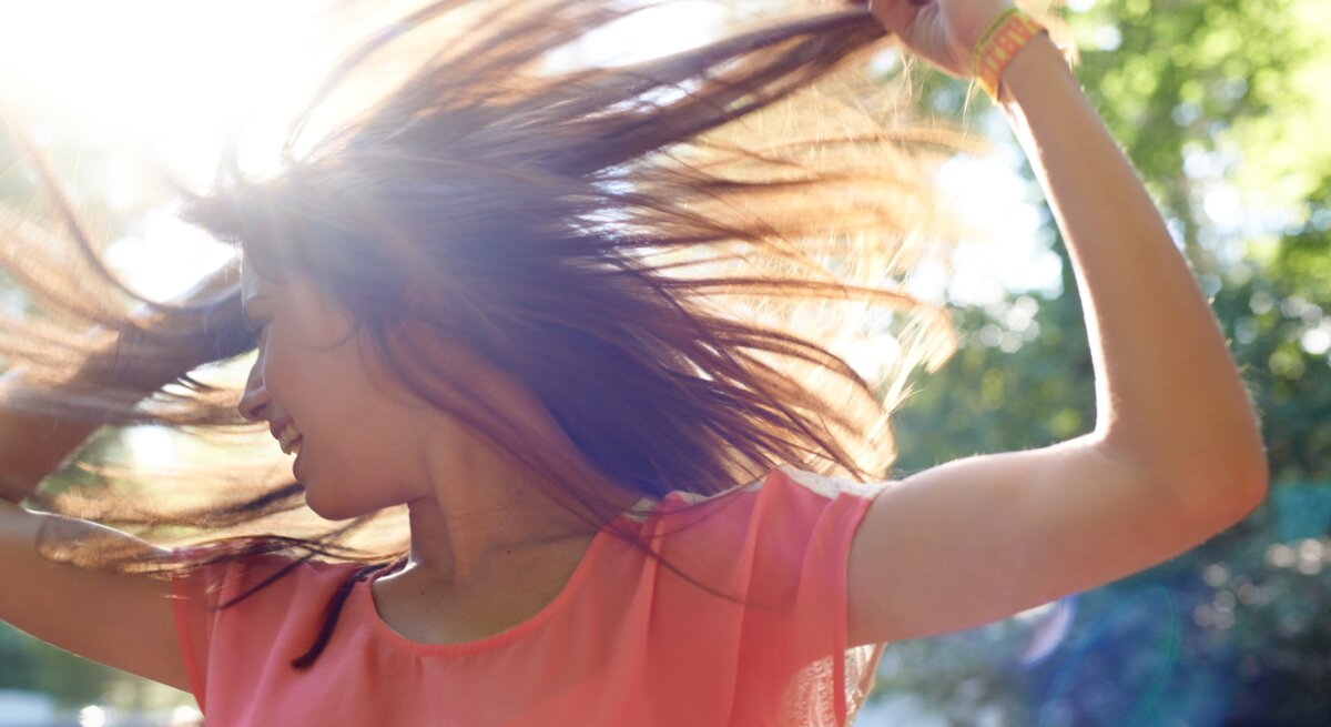 Eine junge Frau mit langen braunen Haaren tanzt lachend in der Sonne, ihr rosafarbenes Oberteil leuchtet im Gegenlicht.
