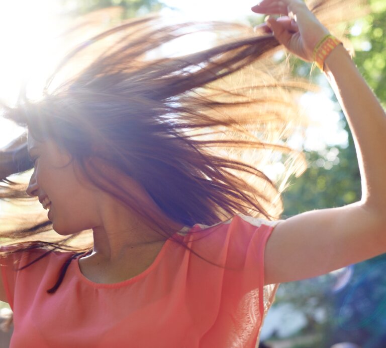 Eine junge Frau mit langen braunen Haaren tanzt lachend in der Sonne, ihr rosafarbenes Oberteil leuchtet im Gegenlicht. | © iStock/PeopleImages