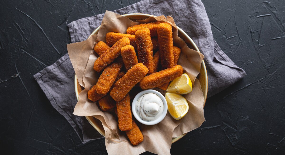 Fischstäbchen mit Remoulade und Zitrone in einer mit Papier ausgelegten Schale. Angerichtet ist das Gericht auf dunkler Arbeitsfläche mit einem Geschirrtuch.
