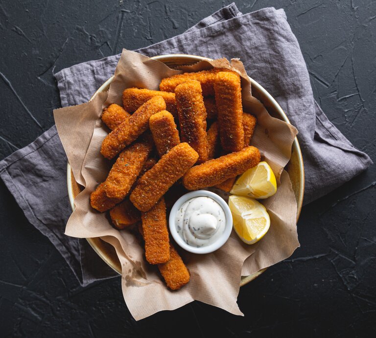 Fischstäbchen mit Remoulade und Zitrone in einer mit Papier ausgelegten Schale. Angerichtet ist das Gericht auf dunkler Arbeitsfläche mit einem Geschirrtuch. | © Getty Images/Rus32