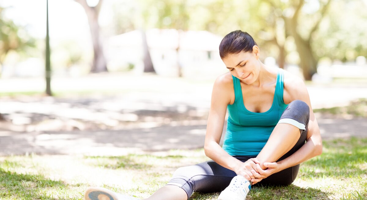 Eine Frau in Sportkleidung sitzt auf dem Gras in einem Park und berührt ihren Fußknöchel, als ob sie Schmerzen hätte. Sie trägt ein türkisfarbenes Tanktop, schwarze Sportleggings und Laufschuhe. Die Szene wirkt sonnig und friedlich, mit Bäumen und einer verschwommenen Umgebung im Hintergrund, was darauf hindeutet, dass sie sich inmitten einer entspannenden Naturumgebung befindet.