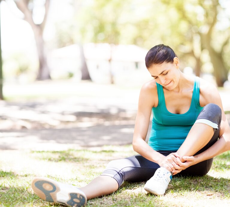 Eine Frau in Sportkleidung sitzt auf dem Gras in einem Park und berührt ihren Fußknöchel, als ob sie Schmerzen hätte. Sie trägt ein türkisfarbenes Tanktop, schwarze Sportleggings und Laufschuhe. Die Szene wirkt sonnig und friedlich, mit Bäumen und einer verschwommenen Umgebung im Hintergrund, was darauf hindeutet, dass sie sich inmitten einer entspannenden Naturumgebung befindet. | © Getty Images/Neustockimages