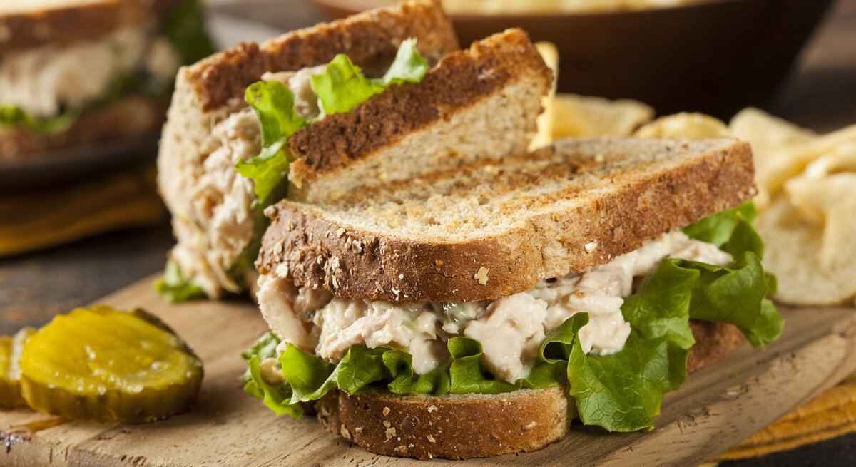 Zwei Thunfisch-Sandwiches auf einem Holzbrett mit einer Gewürzgurkenscheibe und unscharfem weiteren Essen im Hintergrund.