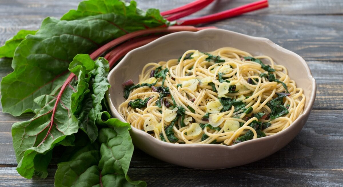 Pasta mit Mangold in cremiger Frischkäse-Soße in beigem Teller auf Holztisch. Daneben bunter Mangold. Draufsicht.