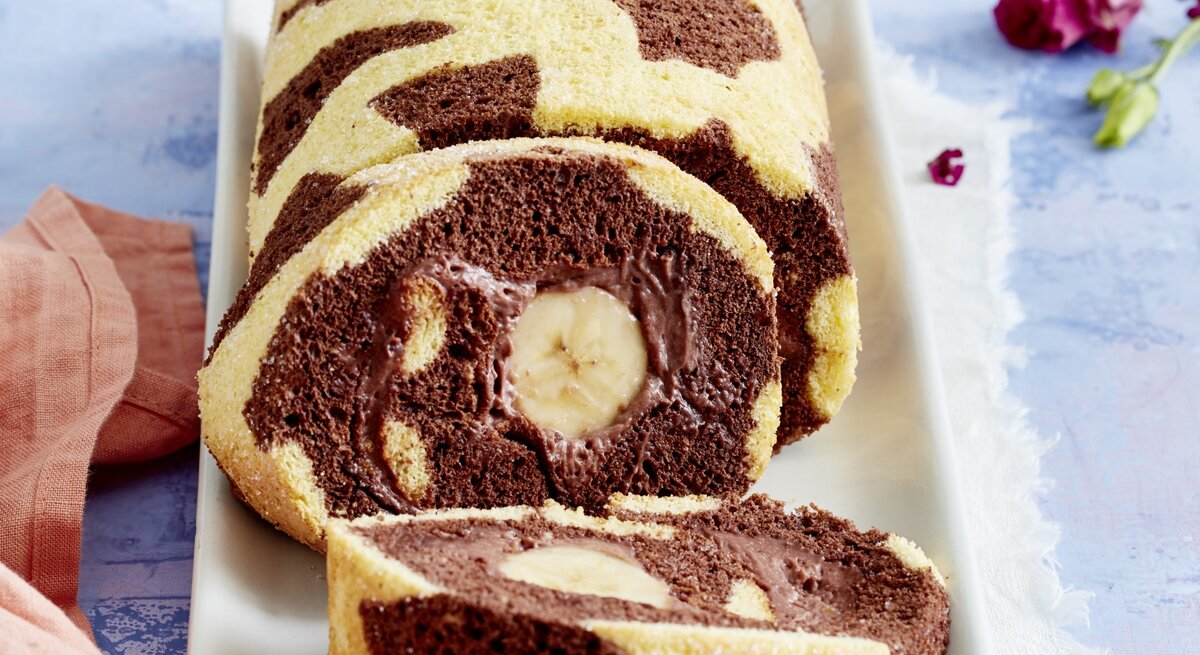 Angeschnittene Giraffen-Bananenrolle mit Schoko-Bananen-Füllung und dunklen Punkten auf hellem Teig. Untergrund hellblau. 