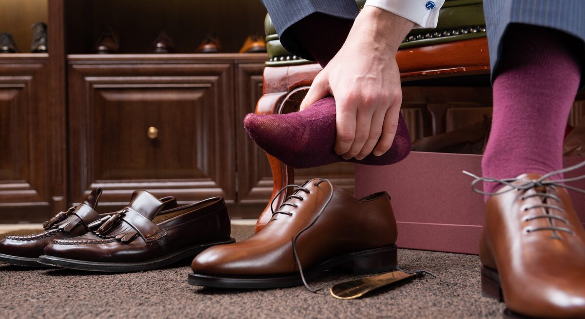 Ein Mann sitzt und zieht einen burgunderroten Socken an seinem Fuß aus, um ihn offenbar gegen ein neues Paar brauner Lederschuhe zu tauschen, die neben ihm stehen. Im Hintergrund sind ein Schuhkarton und weitere Paare von hochwertigen Herrenschuhen auf einem Holzregal zu sehen.