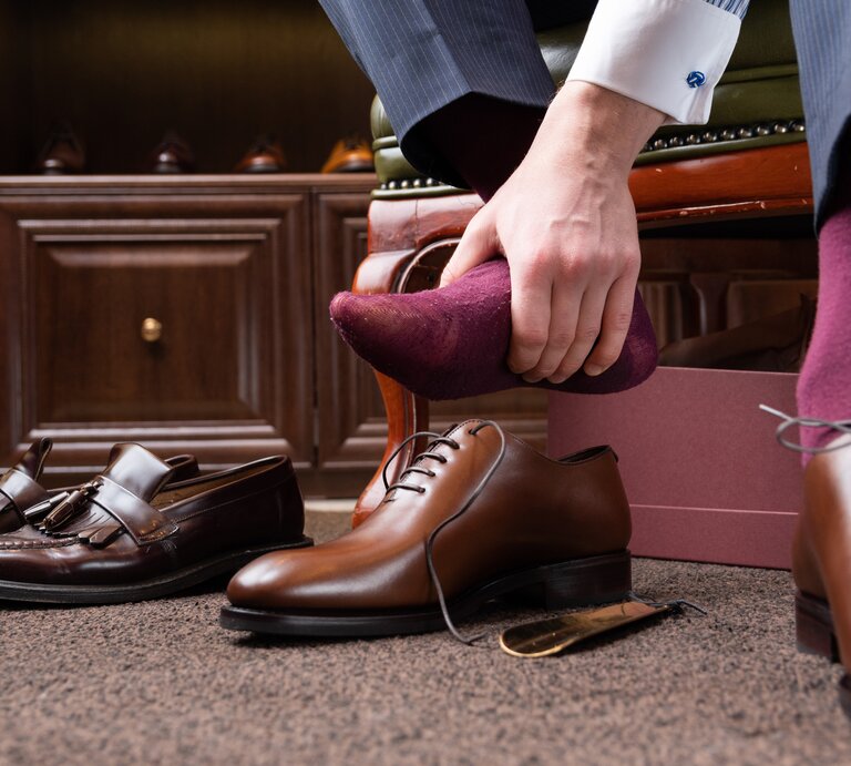 Ein Mann sitzt und zieht einen burgunderroten Socken an seinem Fuß aus, um ihn offenbar gegen ein neues Paar brauner Lederschuhe zu tauschen, die neben ihm stehen. Im Hintergrund sind ein Schuhkarton und weitere Paare von hochwertigen Herrenschuhen auf einem Holzregal zu sehen. | © iStock.com / Sergey Peterman