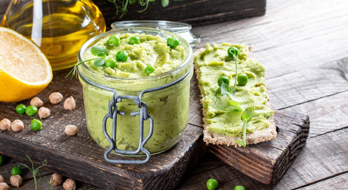 Ein Weckglas mit dem Erbsen-Hummus auf einem Holzbrett mit Zitrone und Olivenöl.