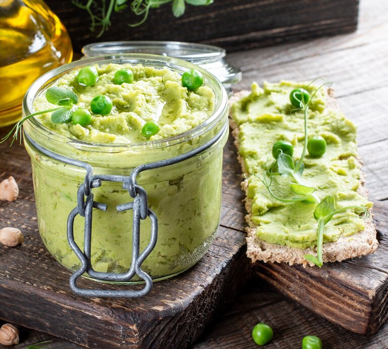 Ein Weckglas mit dem Erbsen-Hummus auf einem Holzbrett mit Zitrone und Olivenöl. | © Shutterstock/ Ahanov Michael