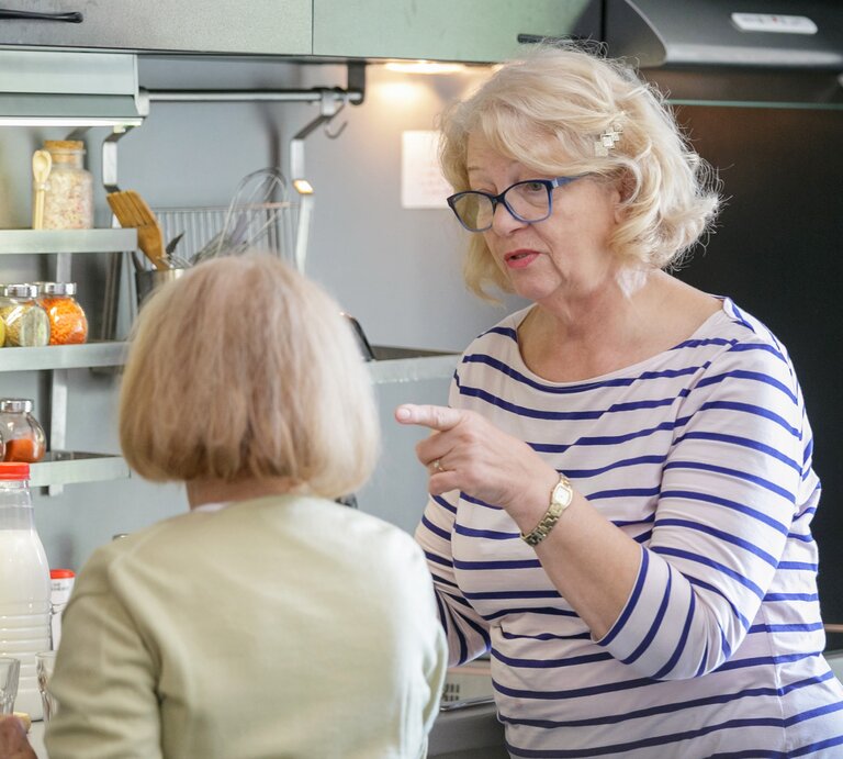 Zwei ältere Frauen sind in einer Küche in eine Unterhaltung vertieft. Die Frau im Vordergrund, mit blonden Haaren und Brille, gestikuliert mit ihrem Finger, was auf eine ernsthafte Diskussion oder Erklärung hinweist. | © Getty Images /	zoranm