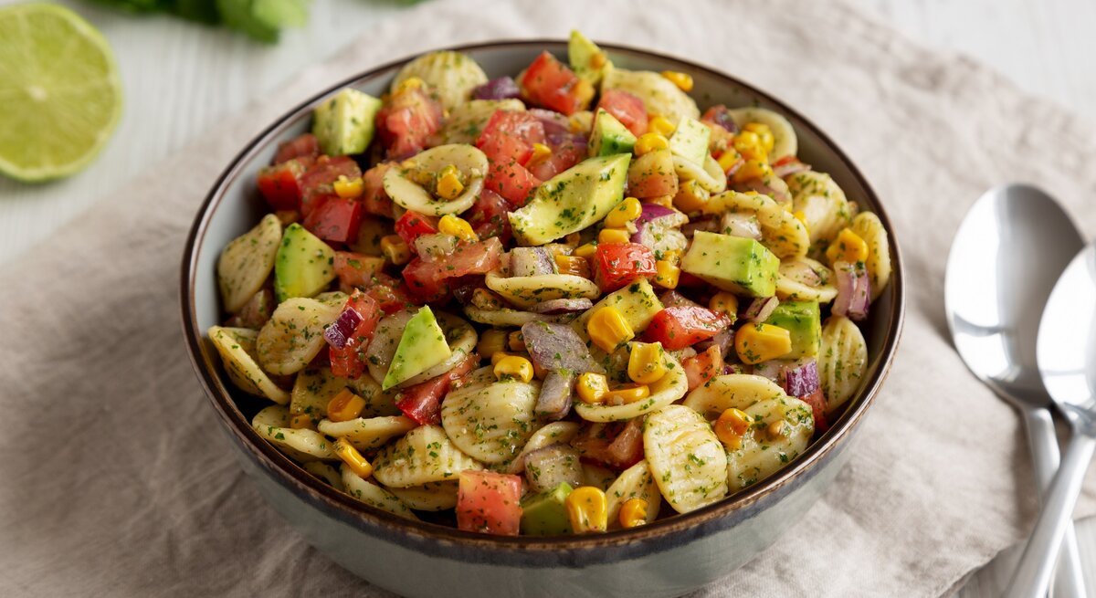 Eine Schüssel Nudelsalat mit Mais und Avocado, außerdem Paprika, drumherum frische Zutaten wie Kräuter und eine halbe Limette, außerdem 2 Löffel.
