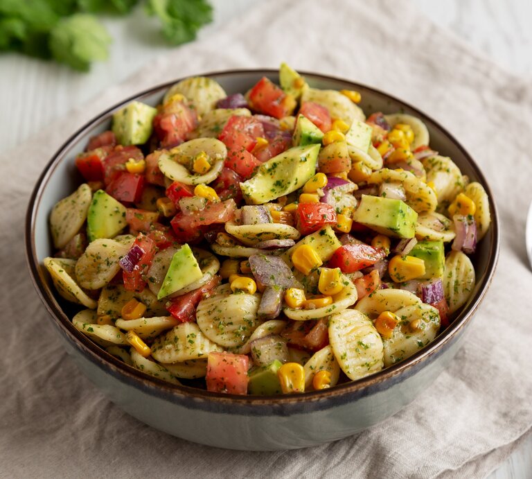 Eine Schüssel Nudelsalat mit Mais und Avocado, außerdem Paprika, drumherum frische Zutaten wie Kräuter und eine halbe Limette, außerdem 2 Löffel. | © Adobe Stock/ Liudmyla