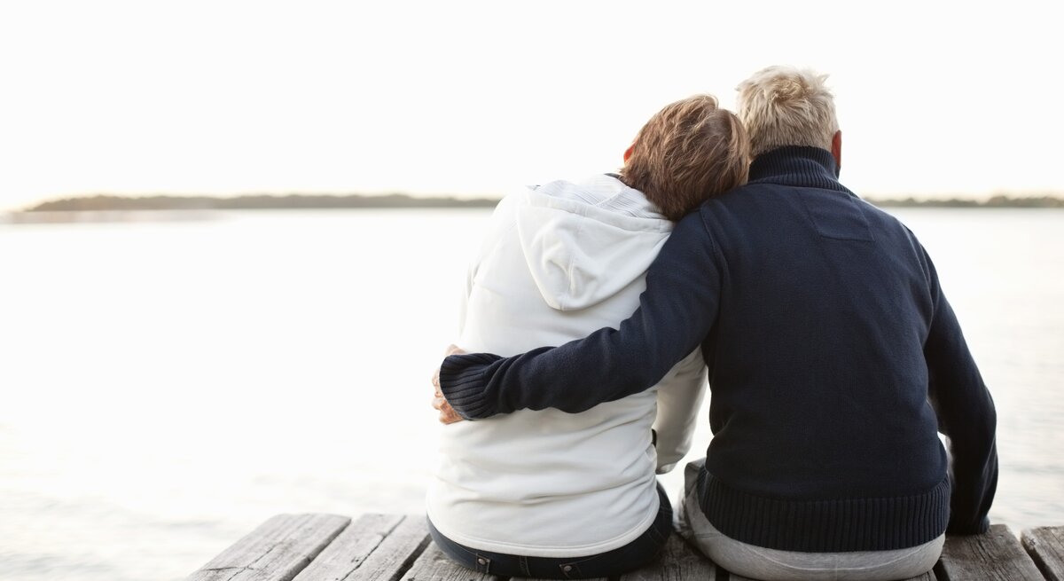 Zwei Personen, die auf einem Steg sitzen und aufs Wasser blicken. Sie sind von hinten zu sehen und tragen warme Kleidung, vermutlich Pullover oder Jacken. Eine Person hat den Arm um die andere gelegt.