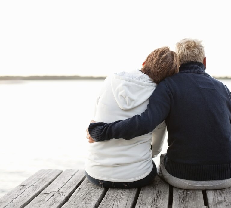 Zwei Personen, die auf einem Steg sitzen und aufs Wasser blicken. Sie sind von hinten zu sehen und tragen warme Kleidung, vermutlich Pullover oder Jacken. Eine Person hat den Arm um die andere gelegt. | © Getty Images / Maskot