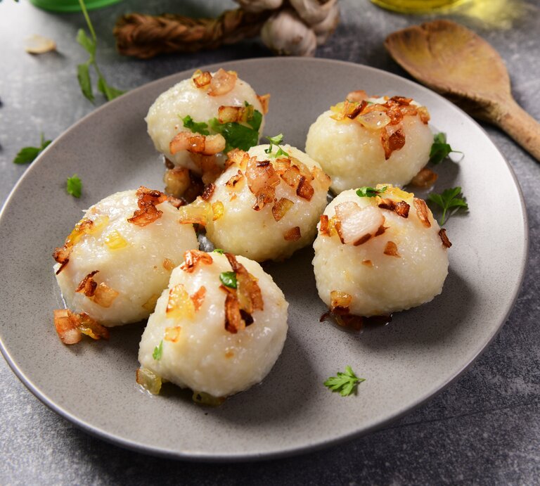 Ein grauer Teller mit den Klößen auf einem steinartigen Untergrund. Darauf geröstete Zwiebeln und daneben ein Holzkochlöffel, Kräuter und Knoblauch. | © Shutterstock/ Piotr Krzeslak