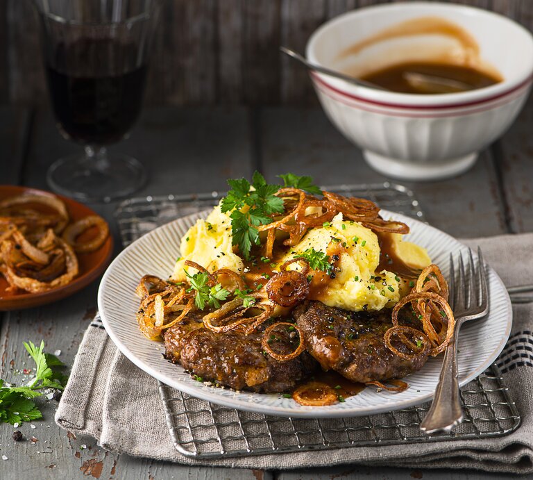 Leckere hausgemachte Hackfleisch-Schnitzel mit Püree und Röstzwiebeln auf einem Teller. Die Schnitzel sind angerichtet mit Bratensoße. Der Teller steht auf Geschirrhandtüchern auf dunklem Holz. | © Stockfood