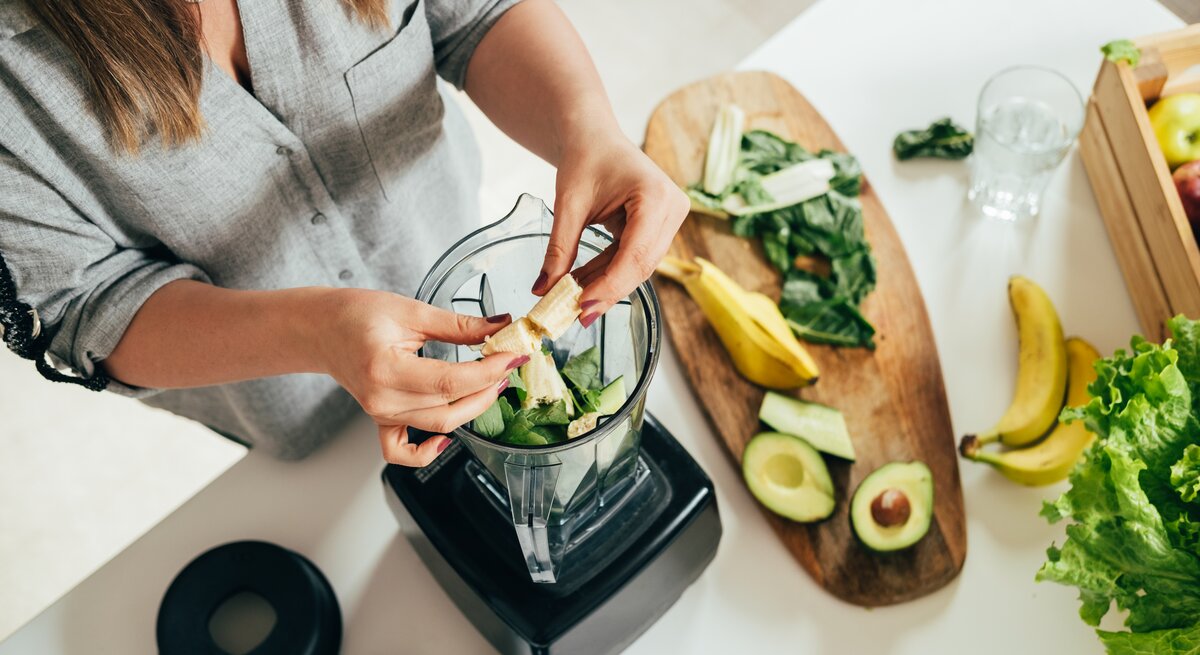 Eine Frau bereitet einen grünen Smoothie vor. Sie gibt frische Zutaten in einen Mixer, darunter Spinatblätter und Bananenstücke. Auf dem Holzbrett neben ihr liegen weitere frische Zutaten wie Avocadohälften, Bananen und Salatblätter. Die Szene spielt in einer hellen, modernen Küche. Der Fokus liegt auf den Händen der Frau und den gesunden Lebensmitteln, was eine Atmosphäre des bewussten Essens und der Gesundheitspflege suggeriert.