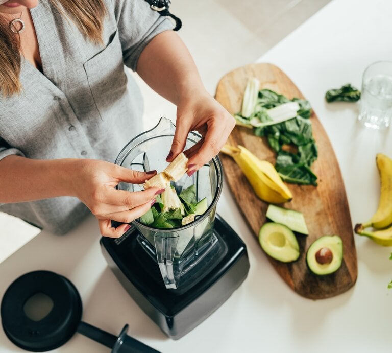 Eine Frau bereitet einen grünen Smoothie vor. Sie gibt frische Zutaten in einen Mixer, darunter Spinatblätter und Bananenstücke. Auf dem Holzbrett neben ihr liegen weitere frische Zutaten wie Avocadohälften, Bananen und Salatblätter. Die Szene spielt in einer hellen, modernen Küche. Der Fokus liegt auf den Händen der Frau und den gesunden Lebensmitteln, was eine Atmosphäre des bewussten Essens und der Gesundheitspflege suggeriert. | © Getty Images/Tijana Simic