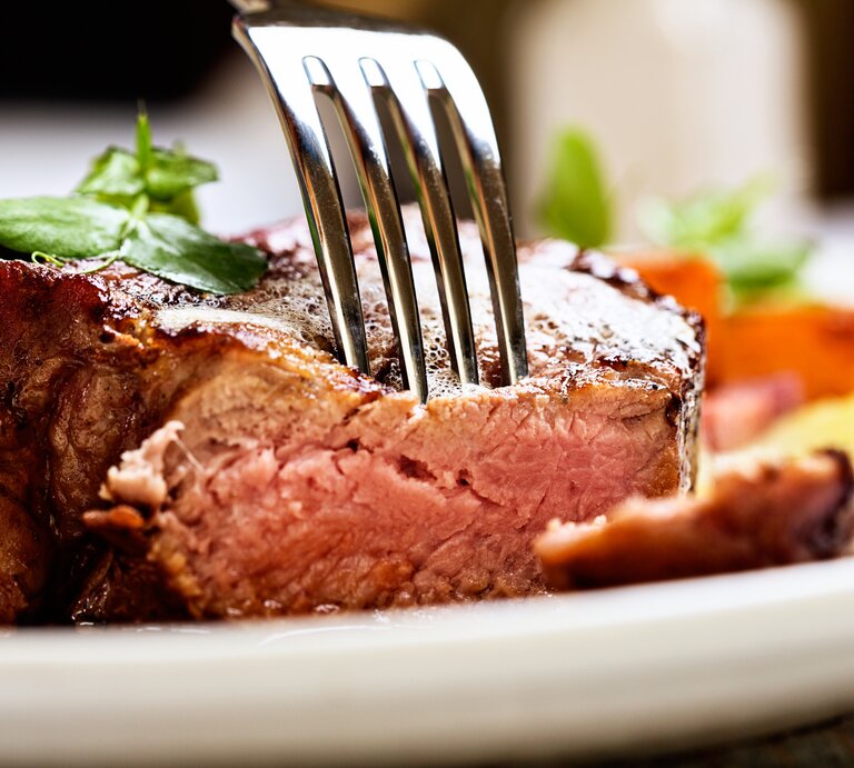 Gabel steckt in einem saftigen Stück Fleisch auf weißem Teller. | © GettyImages/RapidEye