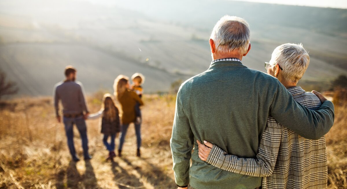 Ein älteres Ehepaar steht Arm in Arm in der Natur, die beiden schauen auf eine junge Familie vor ihnen