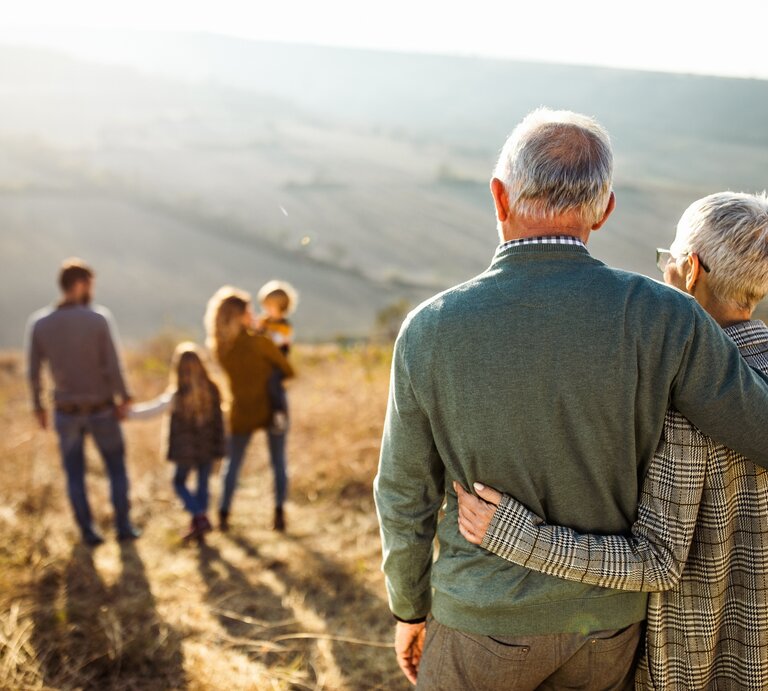 Ein älteres Ehepaar steht Arm in Arm in der Natur, die beiden schauen auf eine junge Familie vor ihnen | © iStock.com/skynesher