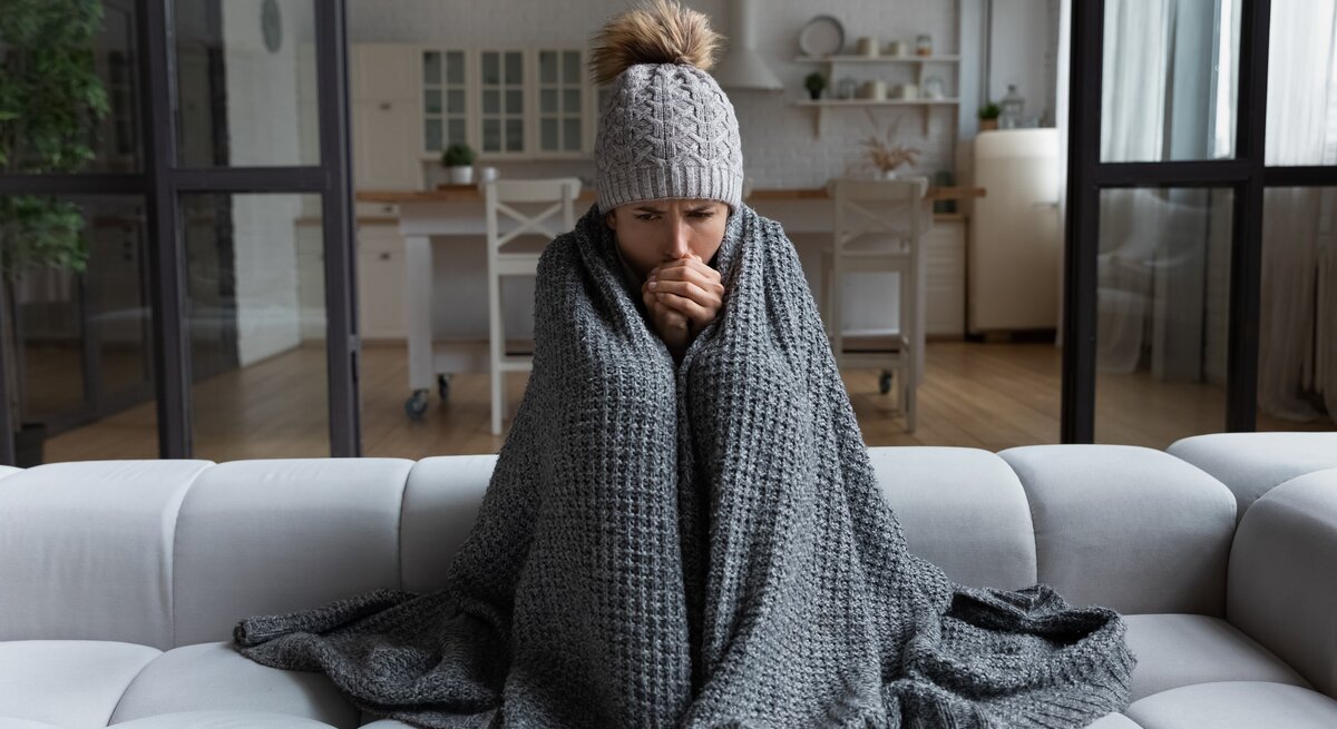 Eine Frau sitzt mit Decke und Mütze auf der Wohnzimmercouch. Sie friert.