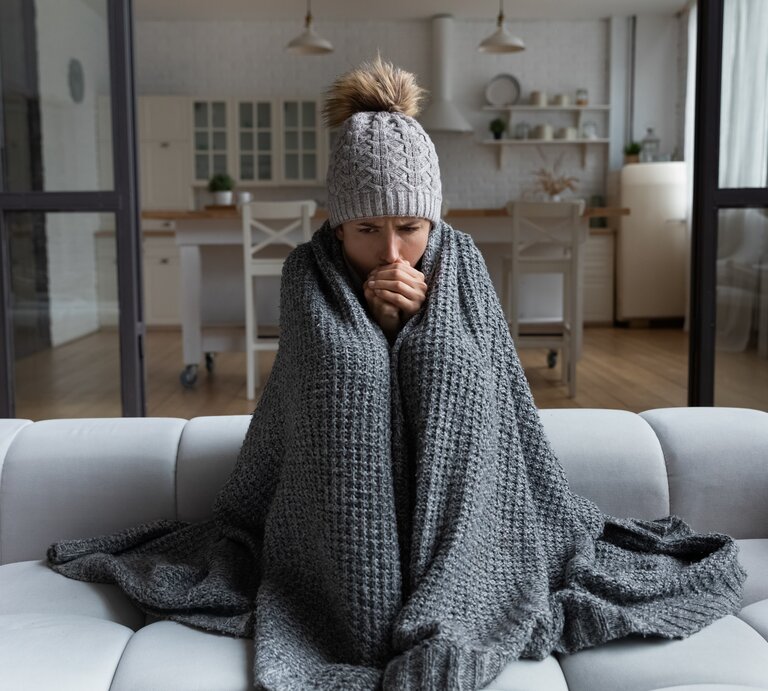 Eine Frau sitzt mit Decke und Mütze auf der Wohnzimmercouch. Sie friert. | © shutterstock/fizkes