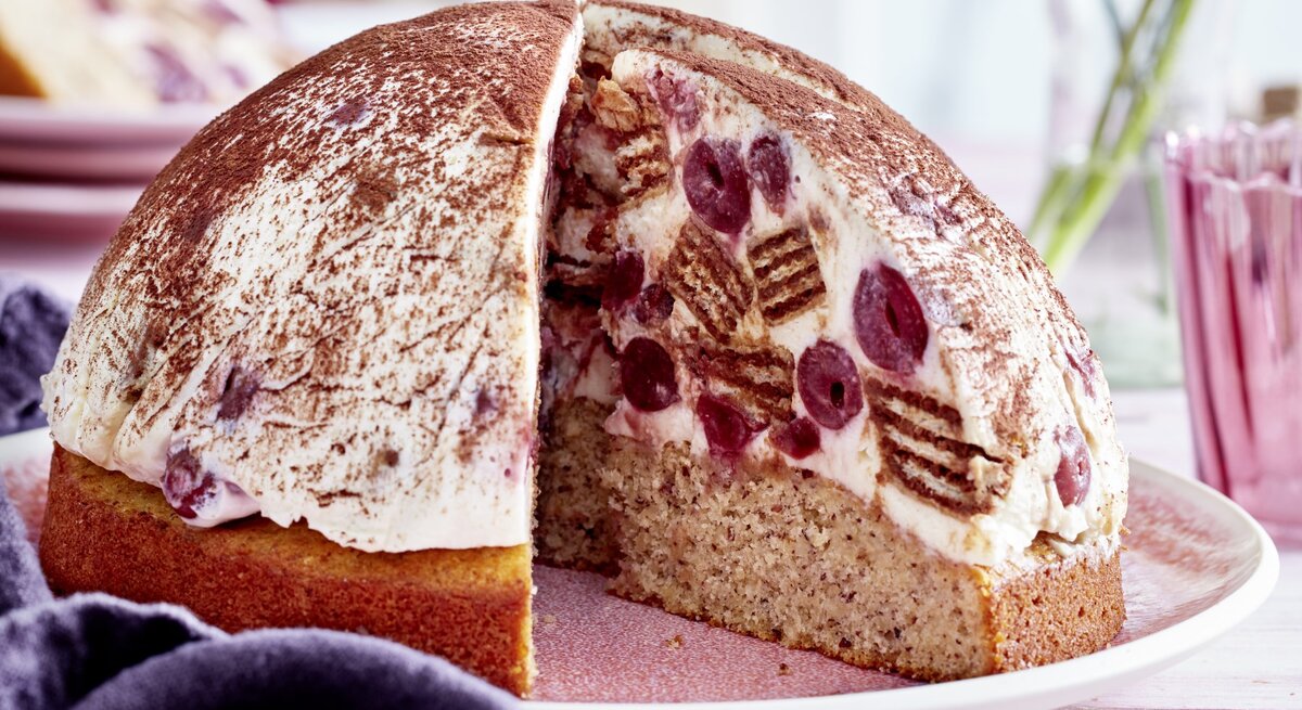 Kugelförmige Tiramisu-Kuppeltorte, gefüllt mit saftigen Kirschen und Waffeln und einem Nussboden, serviert auf einem hellen Teller vor rosarotem Hintergrund. 