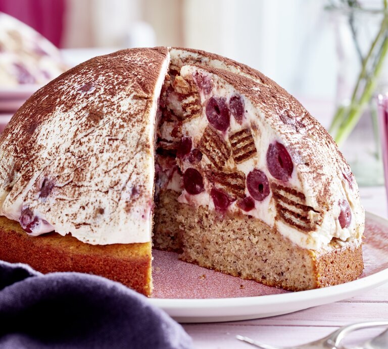 Kugelförmige Tiramisu-Kuppeltorte, gefüllt mit saftigen Kirschen und Waffeln und einem Nussboden, serviert auf einem hellen Teller vor rosarotem Hintergrund.  | © EAT CLUB / Isabell Triemer