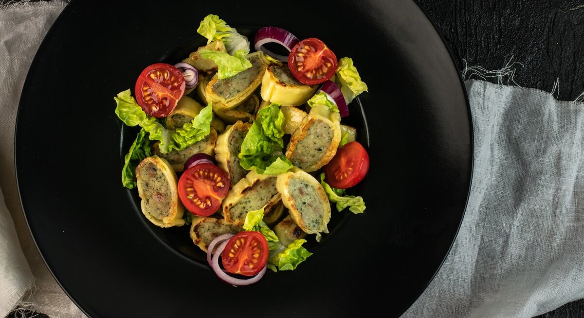 Ein schwarzer Teller mit dem Maultaschensalat auf einem dunklen Tisch mit hellgrauer Tischdecke. Direkt von oben fotografiert.